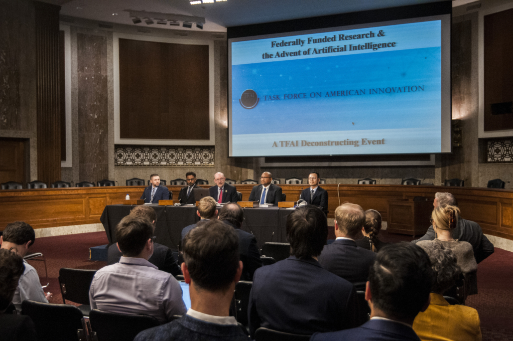 A panel of five experts, with Dan Reed, sitting in the middle, moderating the proceedings of the TFAI Deconstructing AI Senate Briefing. Behind the group is a large screening showing the title of the briefing.
